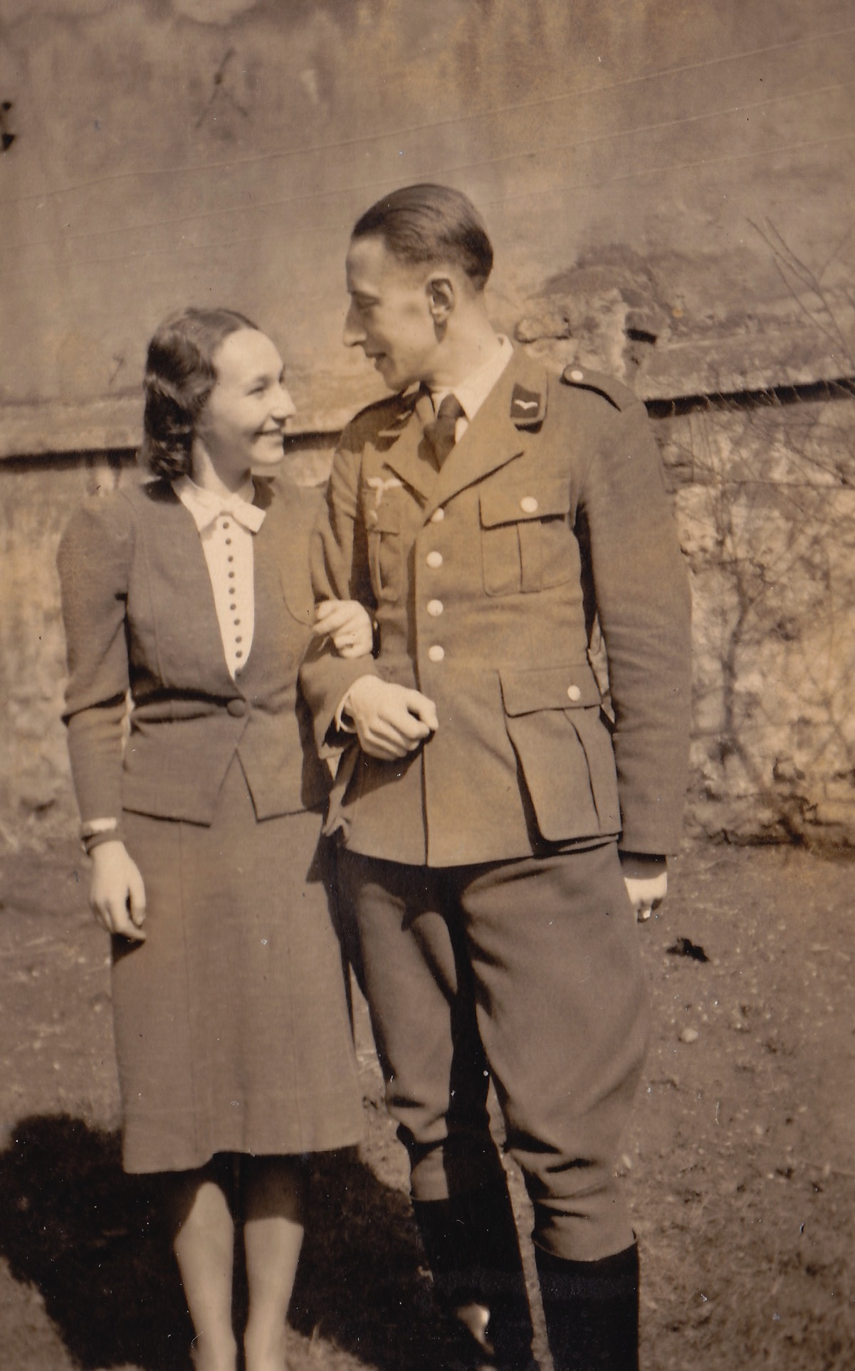 Ernst Willig in German uniform with his wife Margot. Uniform: Luftwaffe – the air force of Nazi Germany
	1.	Collar Tabs:
The distinctive insignia on the collar are characteristic of the Luftwaffe. The specific design suggests a non-commissioned or junior officer rank, although the exact identification is difficult without color.
	2.	Breast Eagle with Swastika:
The emblem on the right breast is the Luftwaffe eagle – a flying eagle clutching a swastika – which was standard on Luftwaffe uniforms. Notably, this eagle was worn on the right side, unlike the Wehrmacht, which placed it on the left.
	3.	Uniform Cut:
The cut and four-pocket design (two breast, two lower) is typical of Luftwaffe tunics (such as the Fliegerbluse or Waffenrock). The shoulder boards appear plain or minimally decorated, suggesting a lower rank.
	4.	Trousers and Boots:
The breeches with high boots are characteristic of military officers or NCOs, often used for formal or field duty.